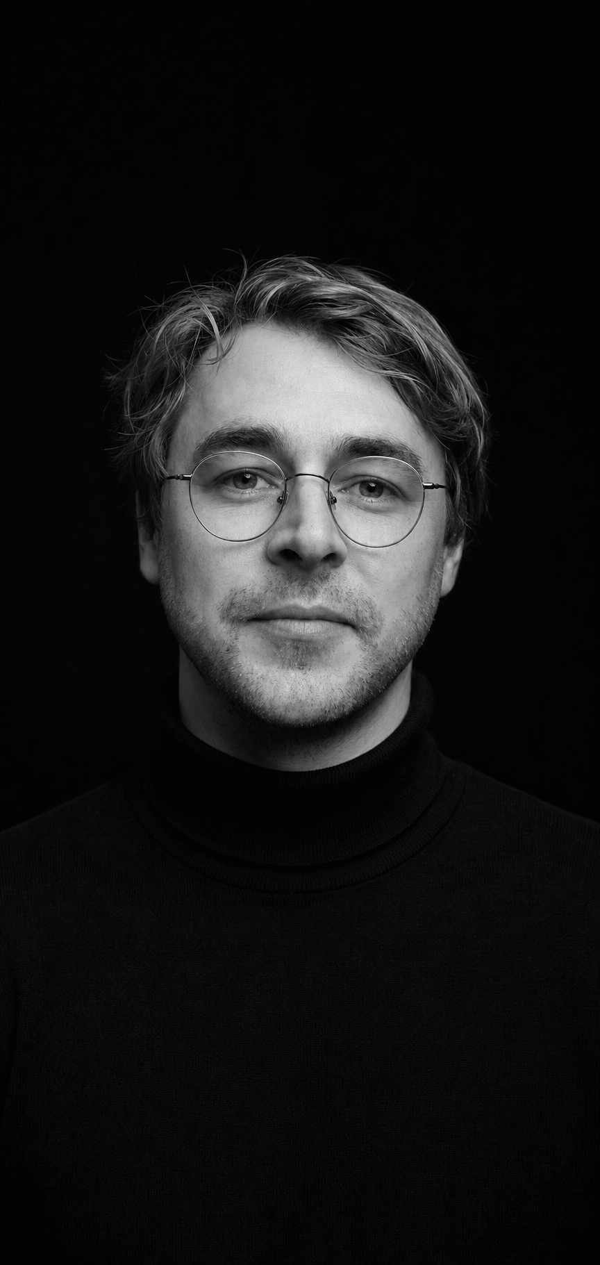 Black-and-white studio portrait of Harry Taylor wearing a black turtleneck and wire-rimmed glasses